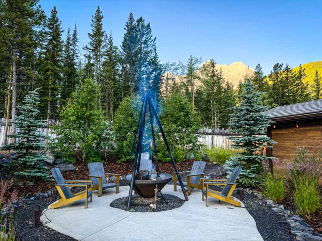 Four wooden chairs surround a tripod fire pit on a circular patio, set outdoors with trees, a wooden fence, and mountains in the background.