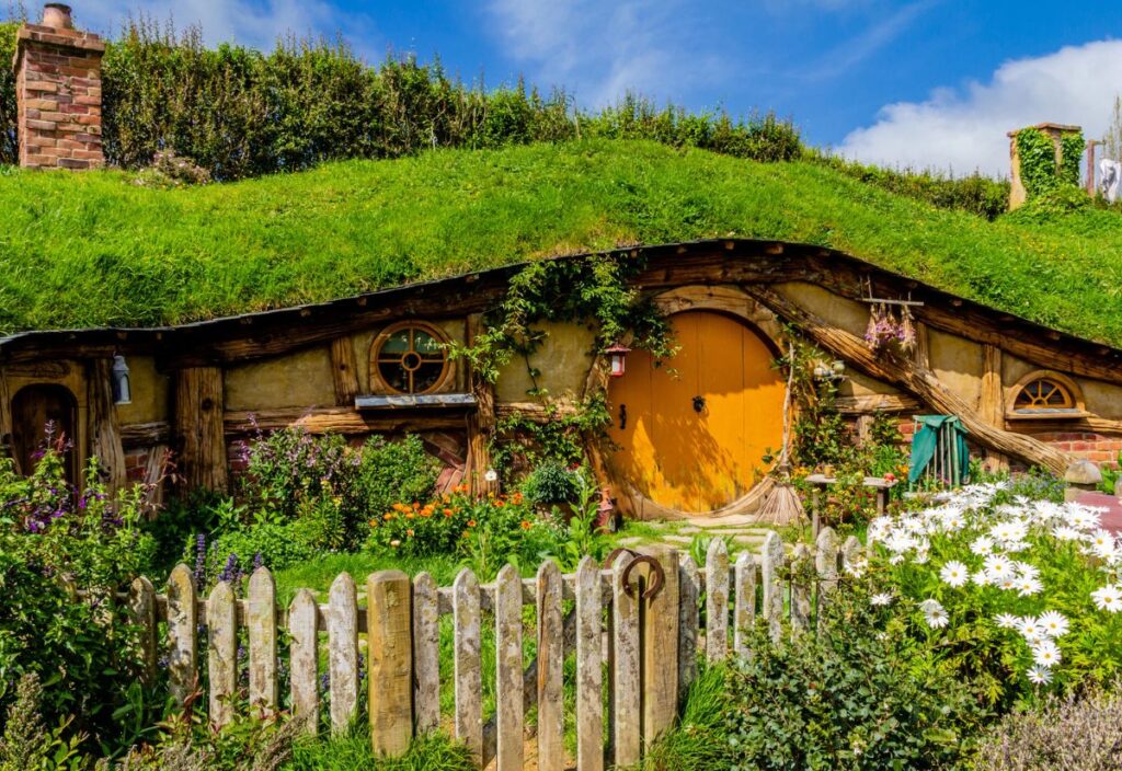 A quaint house with a round yellow door, set into a grassy hillside and surrounded by flowers, evokes the charm of international Hobbit Day beneath a blue sky and behind a wooden picket fence.