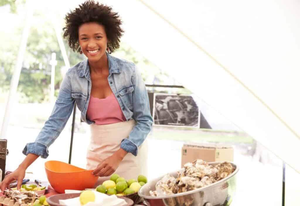 A woman wearing a denim jacket and apron smiles while preparing food at an outdoor table with oysters, limes, and a large orange bowl.