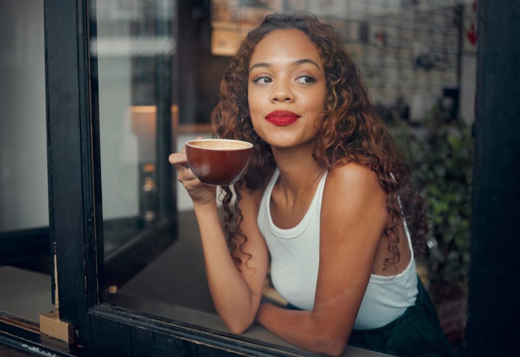 A woman with curly hair in a white tank top holds a cup of coffee while sitting by a window, looking outside thoughtfully, perhaps reflecting on rising coffee prices.