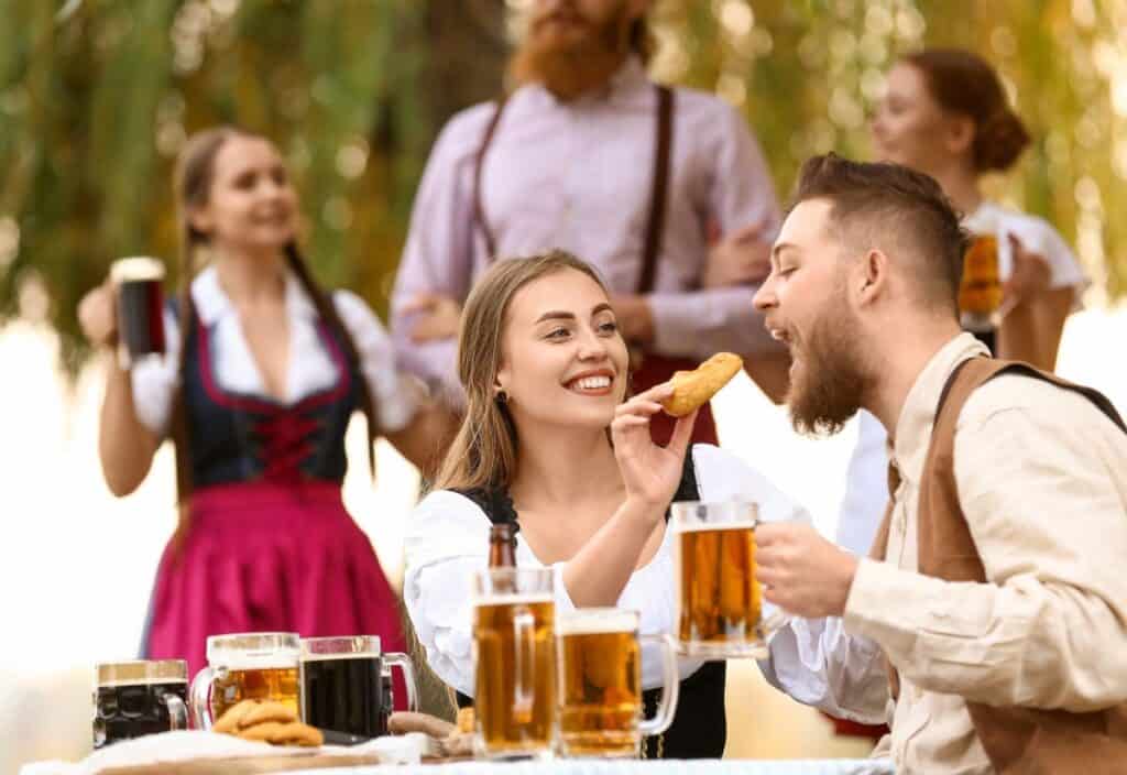 A woman feeds a man a pastry while both hold beer mugs at an outdoor table in the lively spirit often found at festivals across American cities, with others in traditional attire enjoying the festivities in the background.