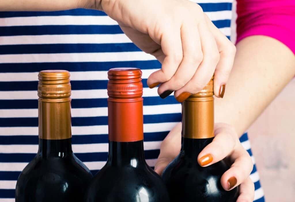 A person in a striped shirt is opening a wine bottle with a screw cap—no corks this time—while two more unopened wine bottles sit beside it.